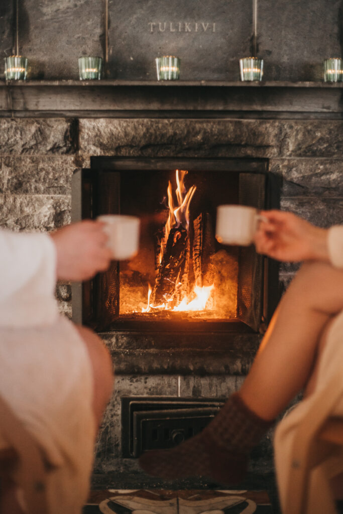 two women holding cups of hot drink sitting near fireplace